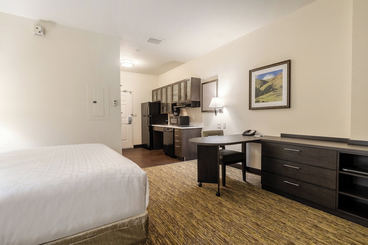 Quarto de hotel com cama, escrivaninha, cadeira, pequena cozinha e quadro pendurado na parede.