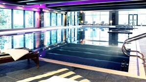 An indoor swimming pool with clear glass walls, a lounge area with a bed and chairs, and a view of the outdoors through large windows. - Village Hotel Farnborough (Farnborough)