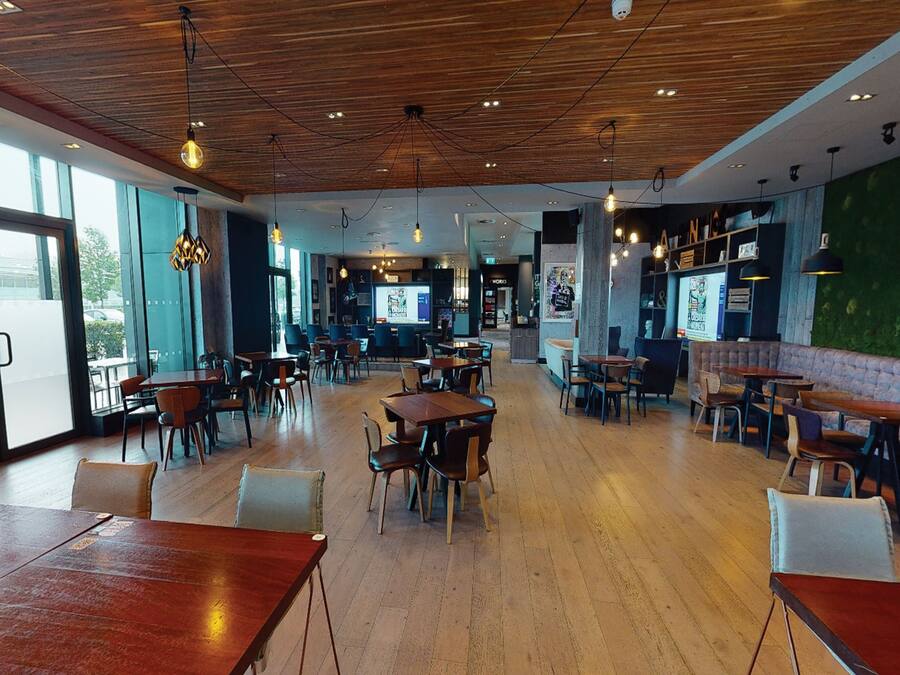 A spacious restaurant with wooden tables and chairs, a bar area, and a green wall with artwork.