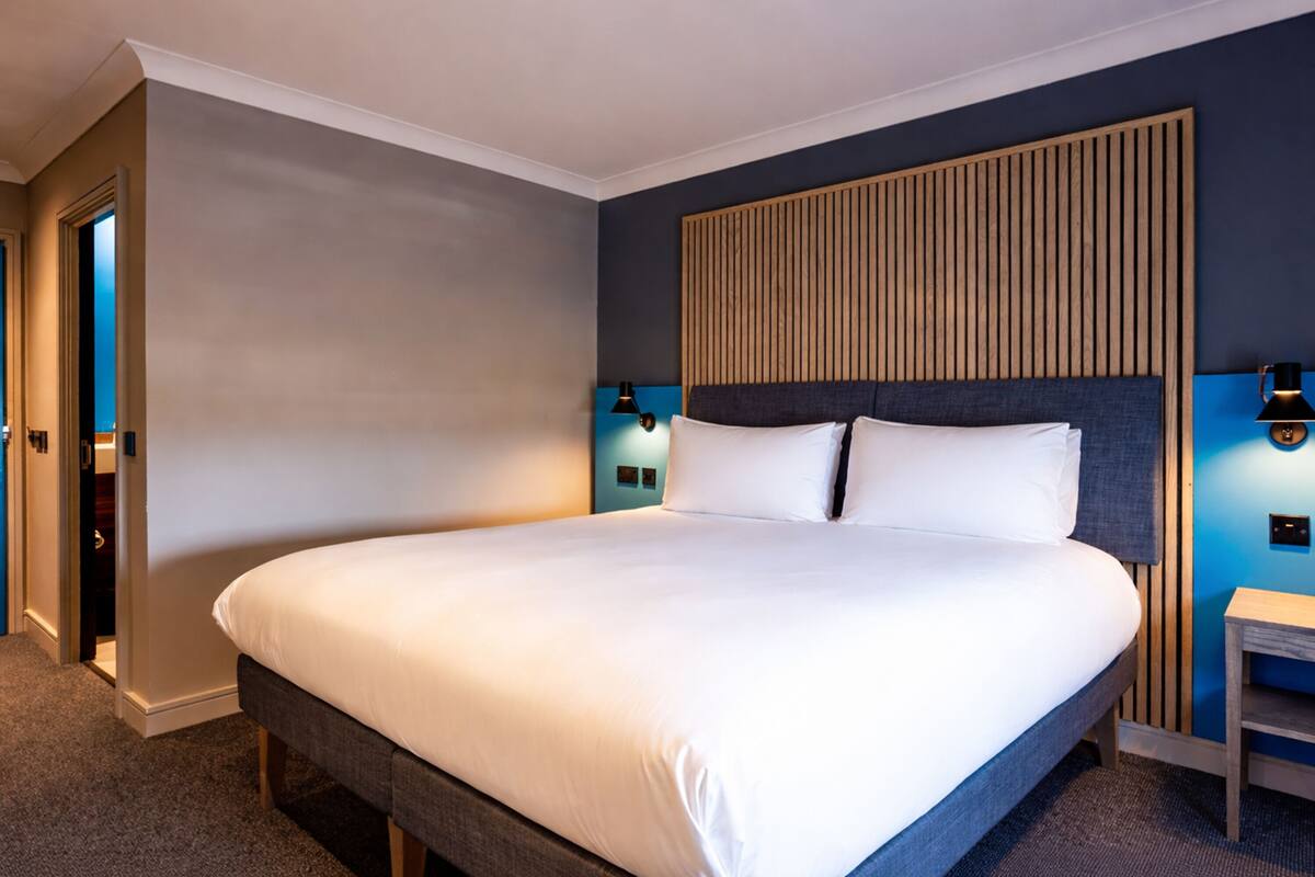 A hotel room with a large bed, a wooden headboard, two bedside lamps, and a small nightstand.