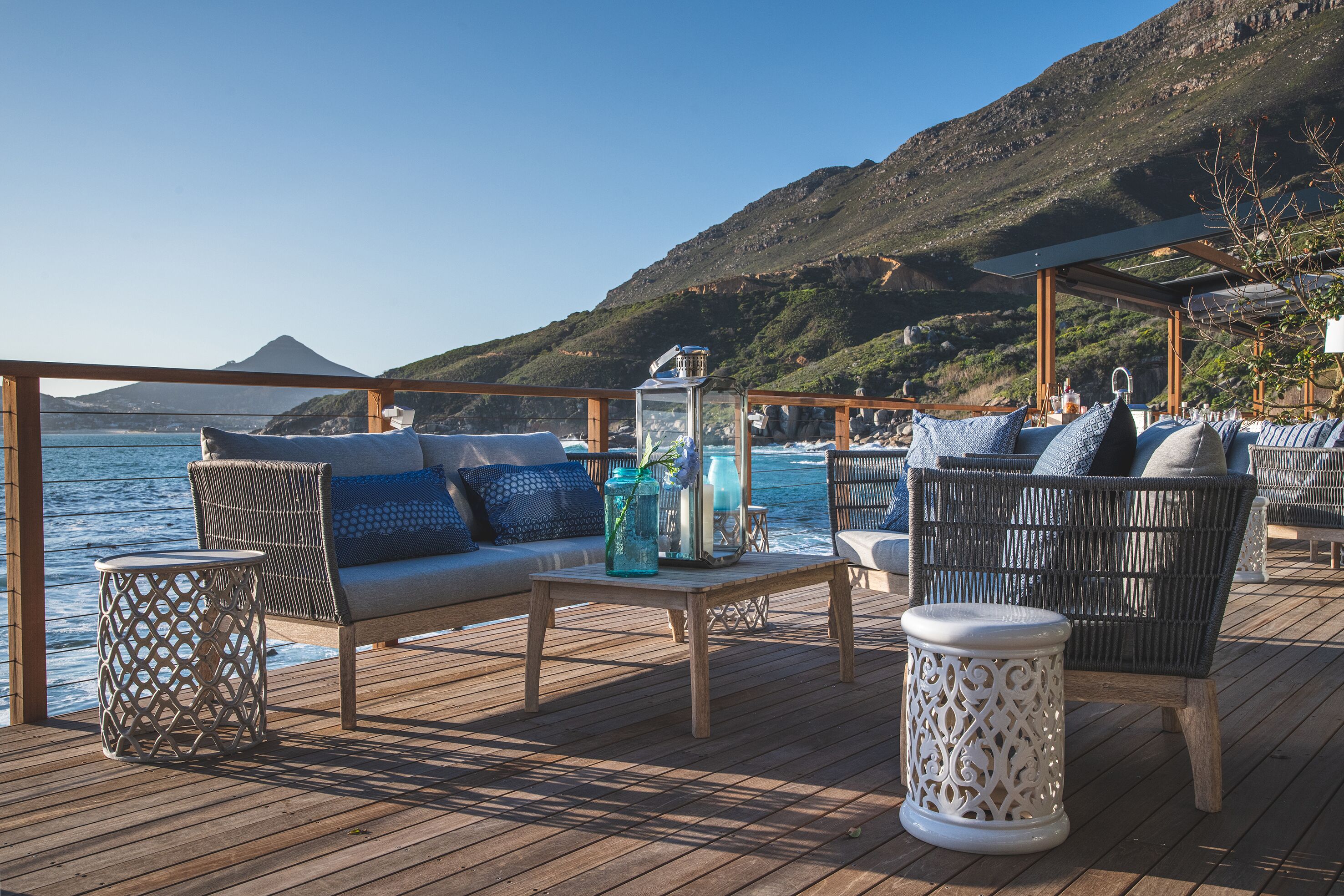 A wooden deck with outdoor seating, including a sofa and chairs, near a body of water with a mountainous backdrop.
