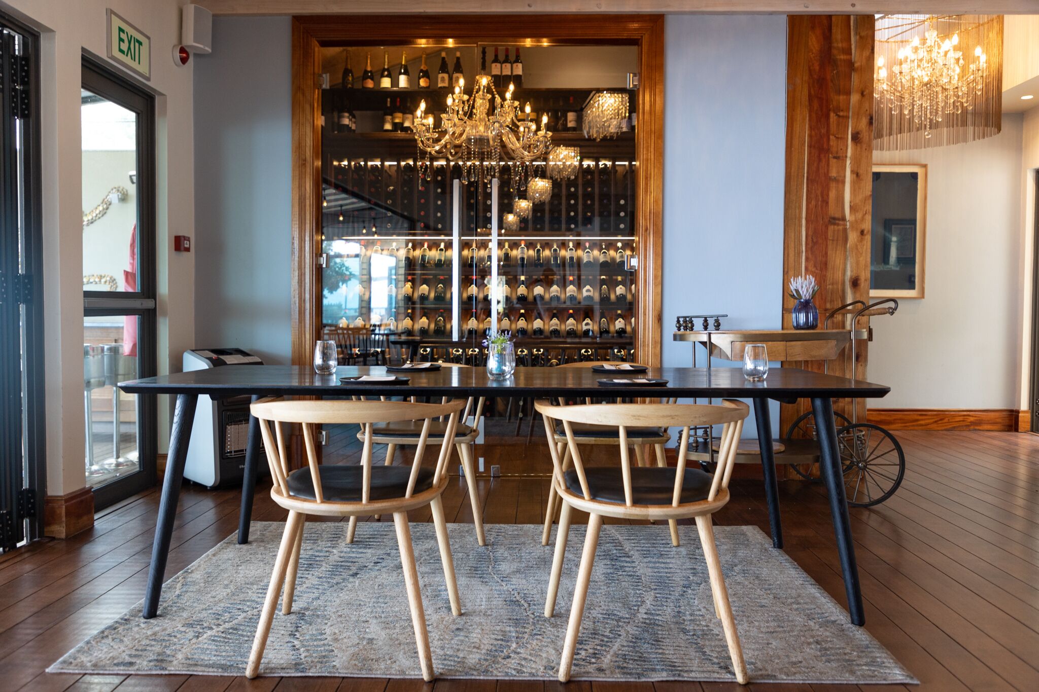 Un comedor con una mesa larga, dos sillas de madera, una araña y una estantería para vinos al fondo.