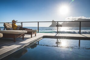 Área de piscina iluminada pelo sol, com espreguiçadeiras e vista para o oceano.