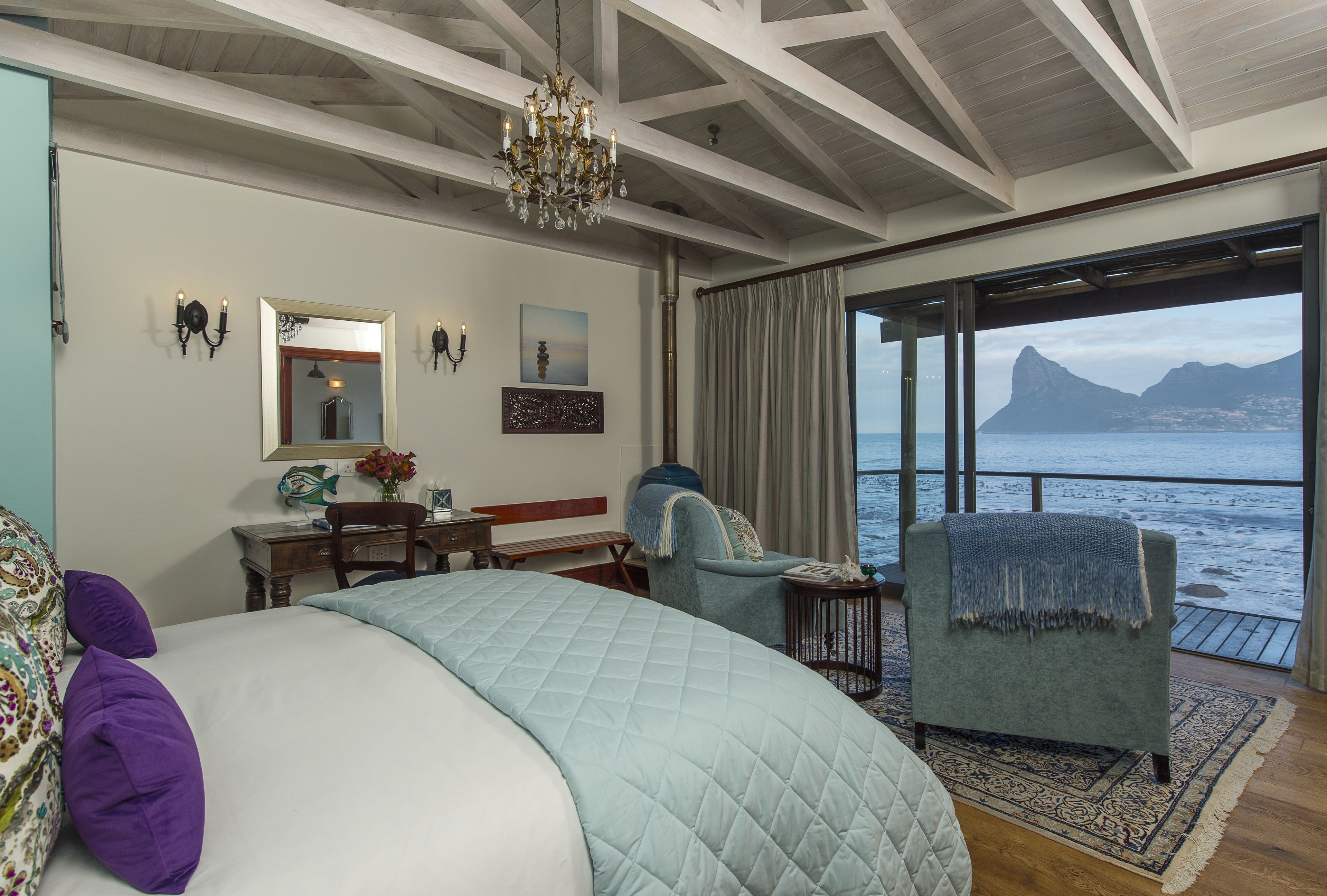 A bedroom with a large bed, a desk, a chair, a chandelier, and a view of the sea.