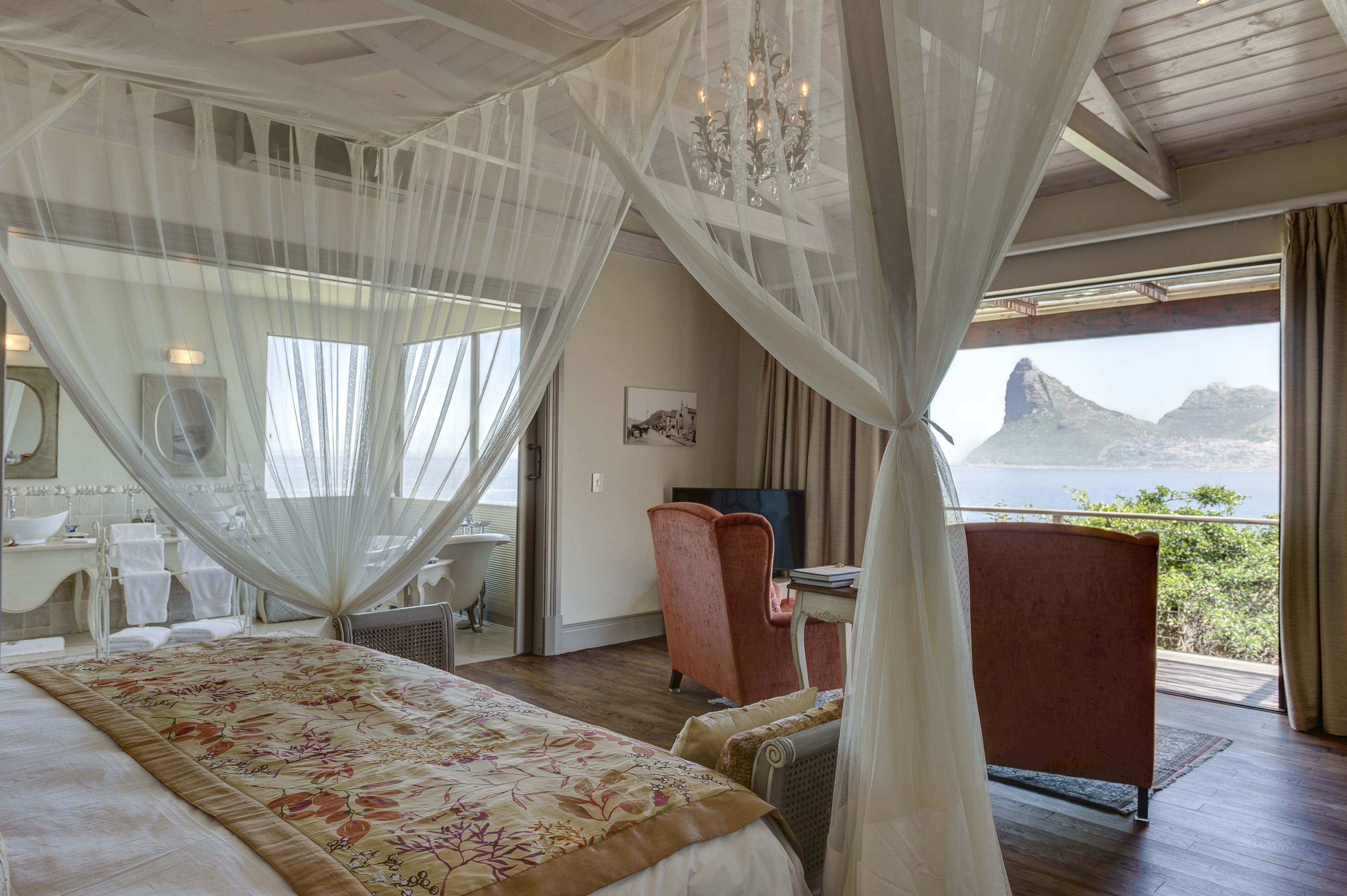 A bedroom with a large bed, a chair, a desk, and a view of a mountain and water.