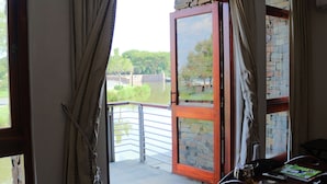 An open balcony with a view of a lake and trees, a table with a glass of drink, and a remote control on the table. - Riboville (Midrand)