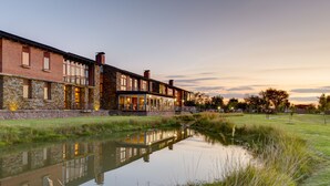 A riverside hotel with stone and brick architecture, a reflective water body, and a grassy area. - Riboville (Midrand)
