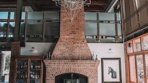 A spacious living room with a high ceiling, exposed wooden beams, a brick fireplace, and a large window. - Riboville (Midrand)