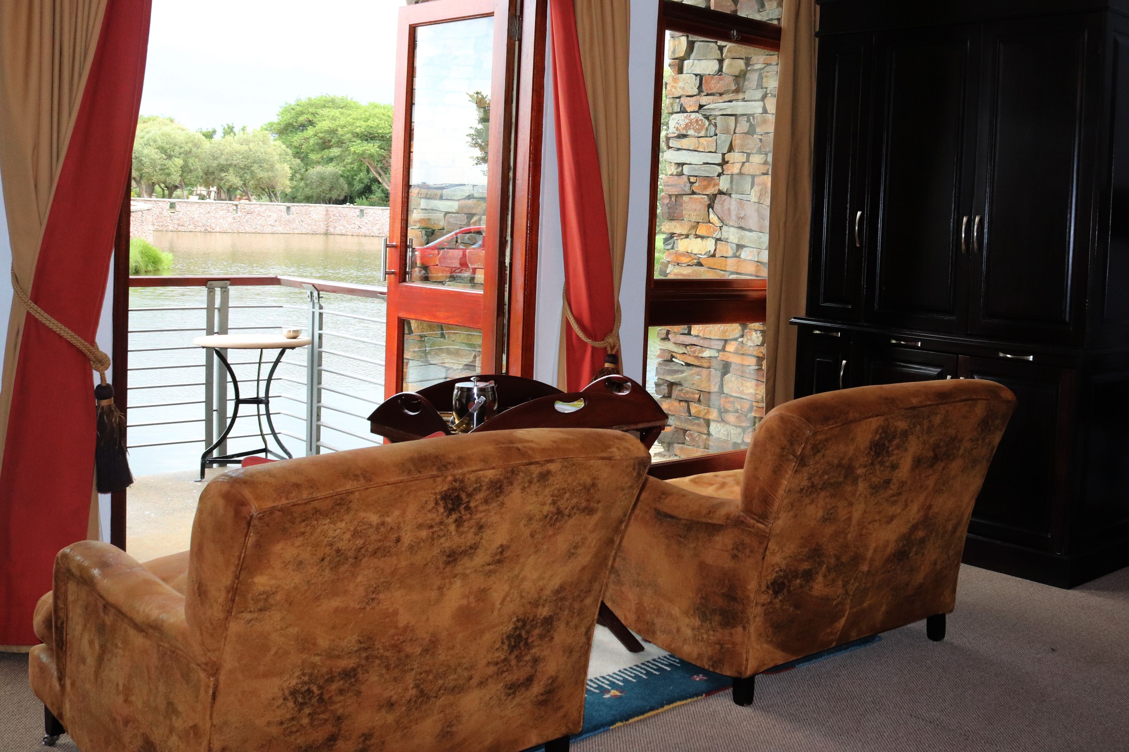 A room with two armchairs, a dining table with chairs, a large black cabinet, and a view of a body of water and trees through a glass door.