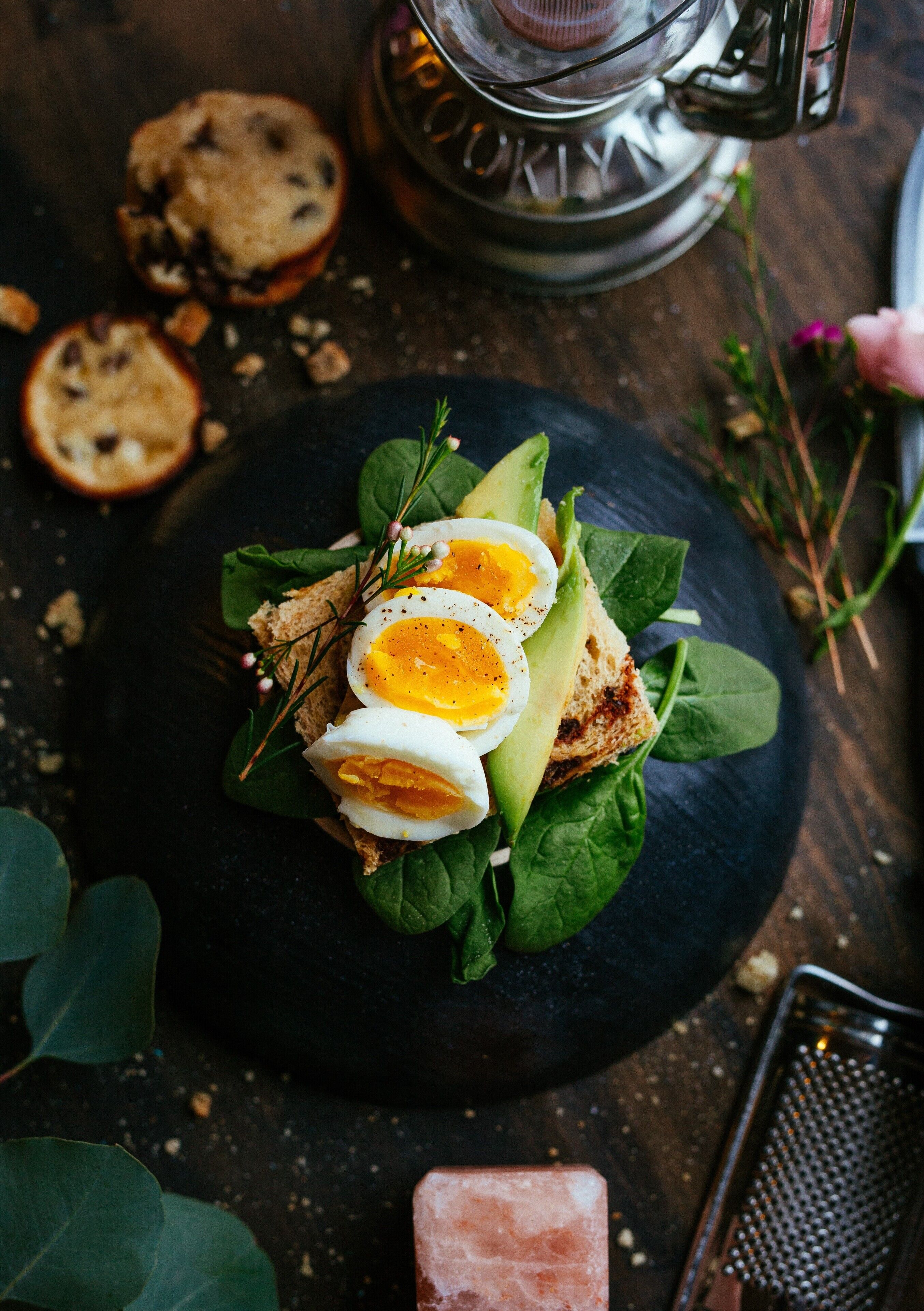 A sandwich on a black plate with spinach leaves, avocado slices, and a boiled egg on top.