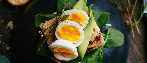 A sandwich on a black plate with spinach leaves, avocado slices, and a boiled egg on top.