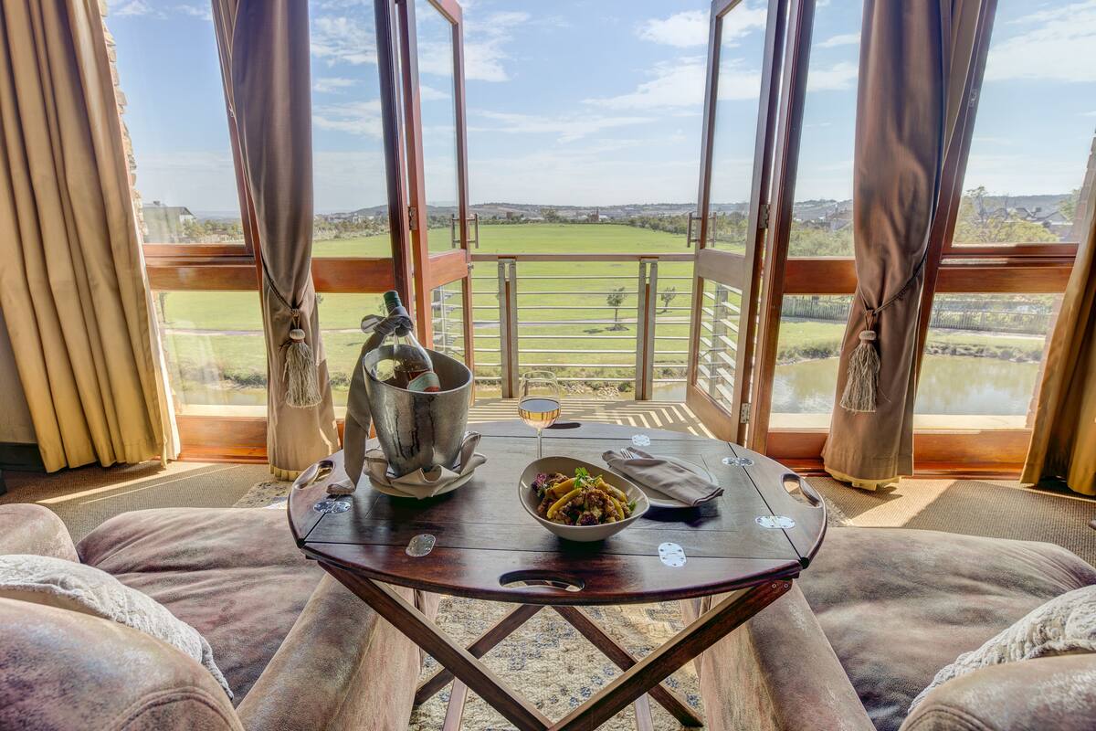 Un comedor con una mesa dispuesta para dos, un plato de comida, una copa de vino y un cubo de metal. La habitación tiene ventanas grandes con cortinas, que ofrecen vistas a un paisaje verde.