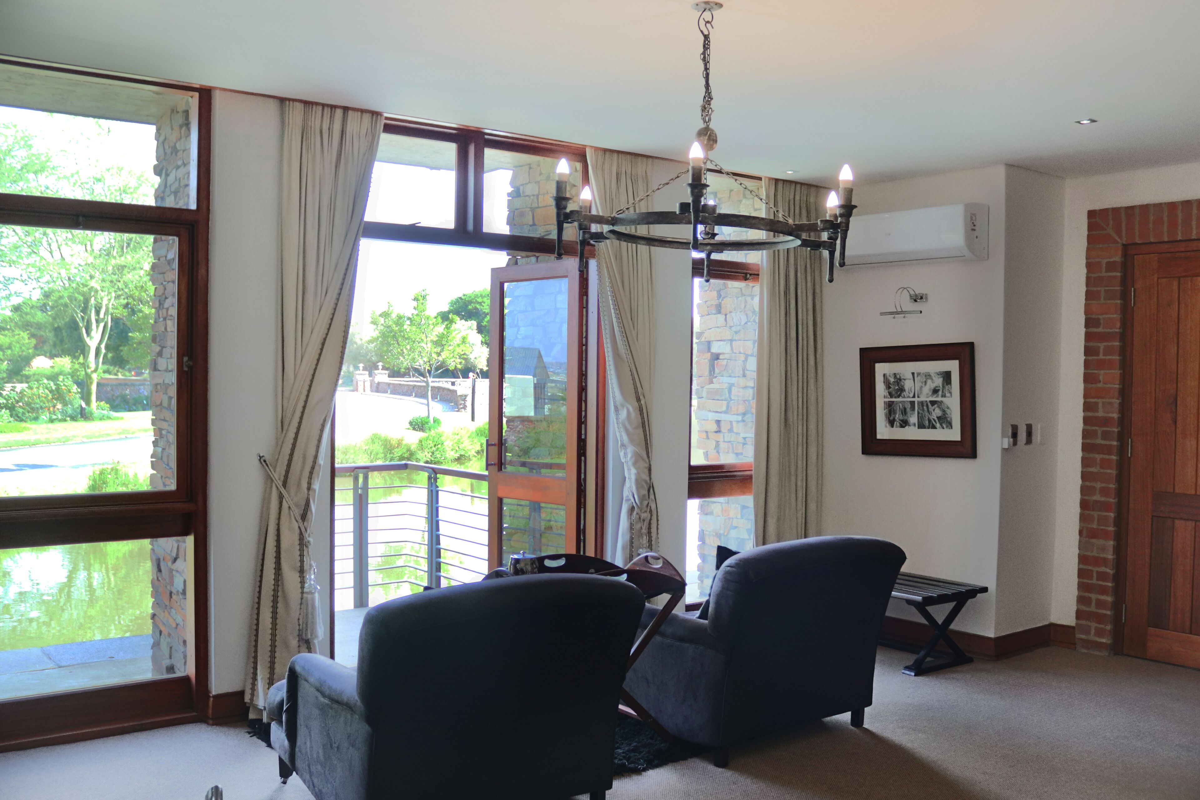 A room with two dark armchairs, a chandelier, a large window with curtains, a view of a garden, and a framed picture on the wall.