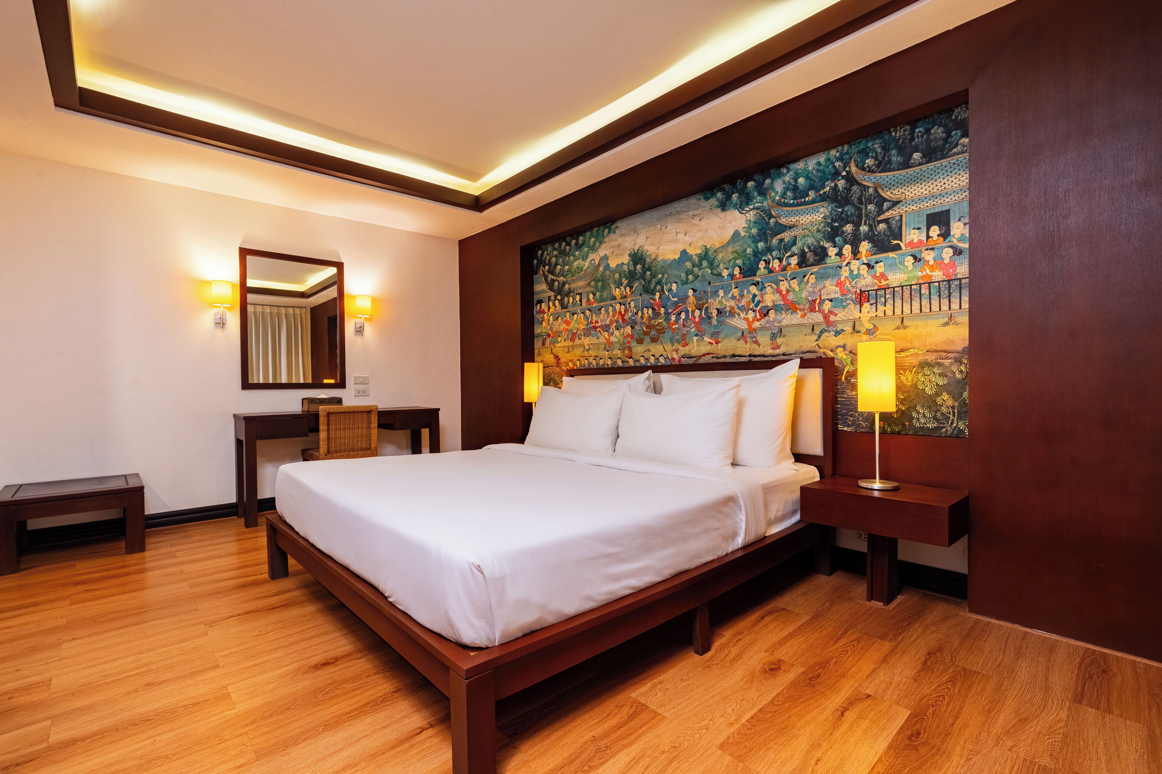 A hotel room with a wooden bed, a desk, a chair, a nightstand, a lamp, a painting, and a mirror.