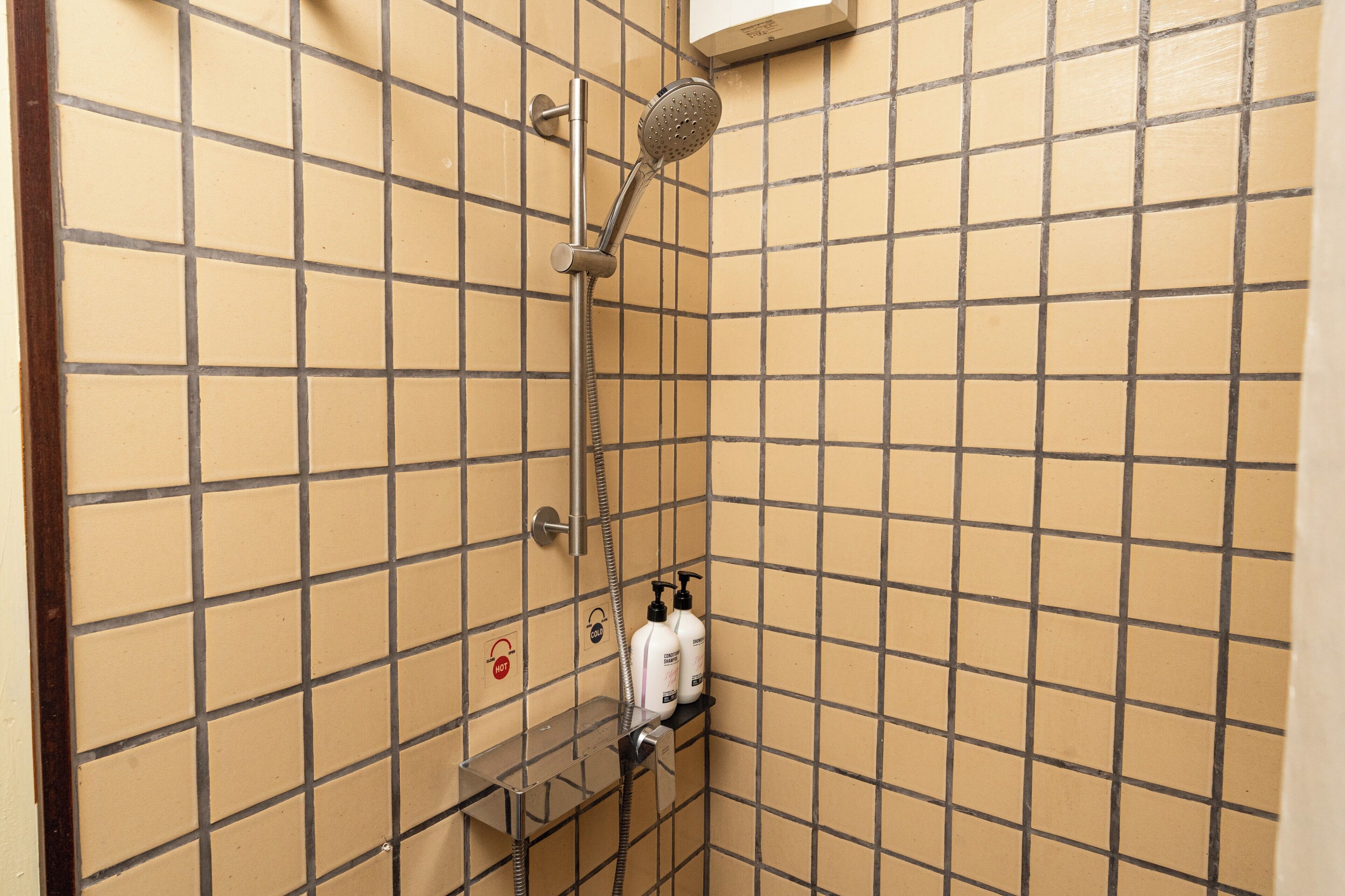 Zona de ducha con azulejos, un cabezal de ducha manual, un grifo empotrado en la pared y un estante con dos botellas.