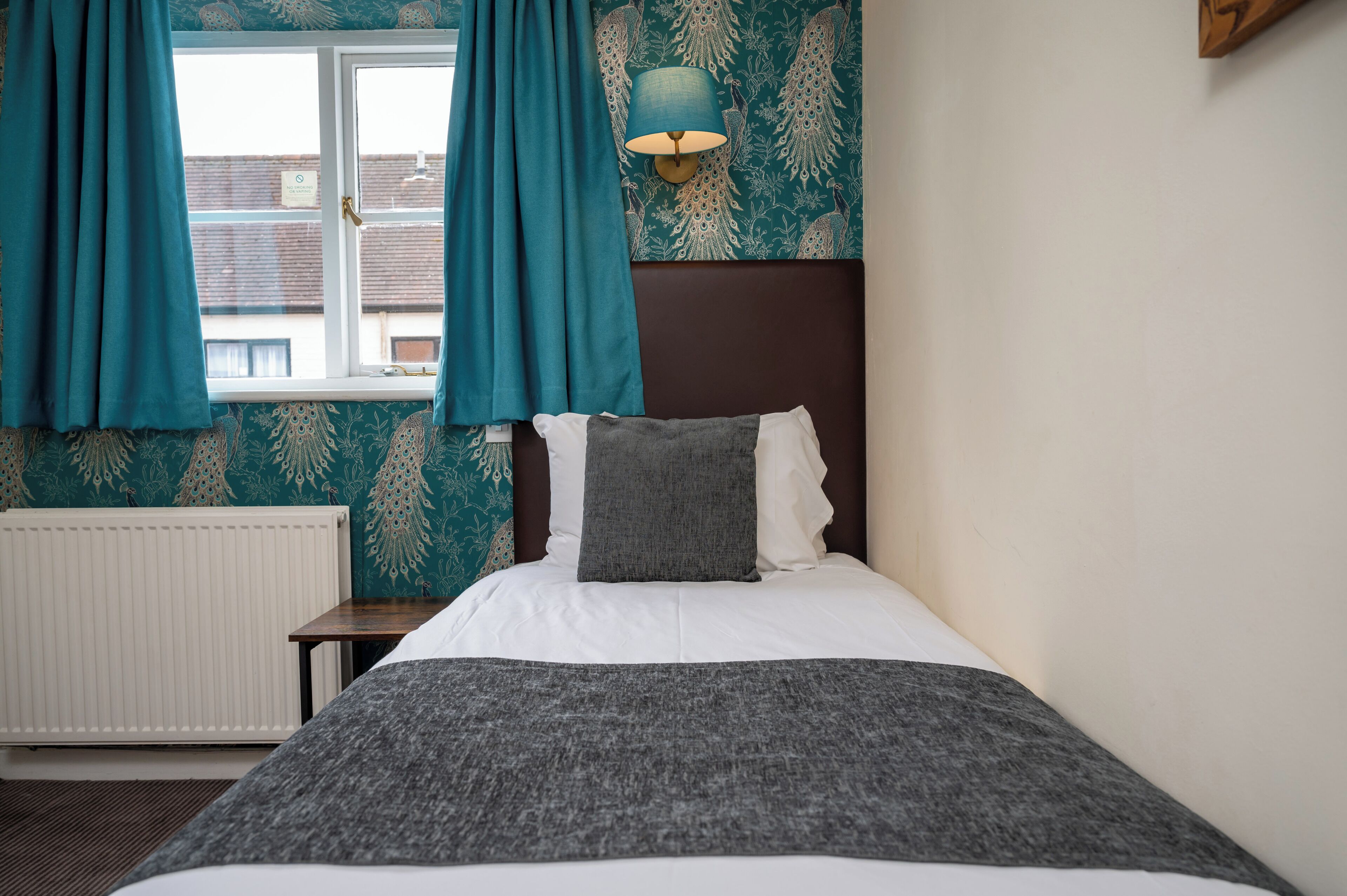 A neatly made bed with a grey blanket and pillows, a bedside table, a wall-mounted lamp, and teal curtains over a window.