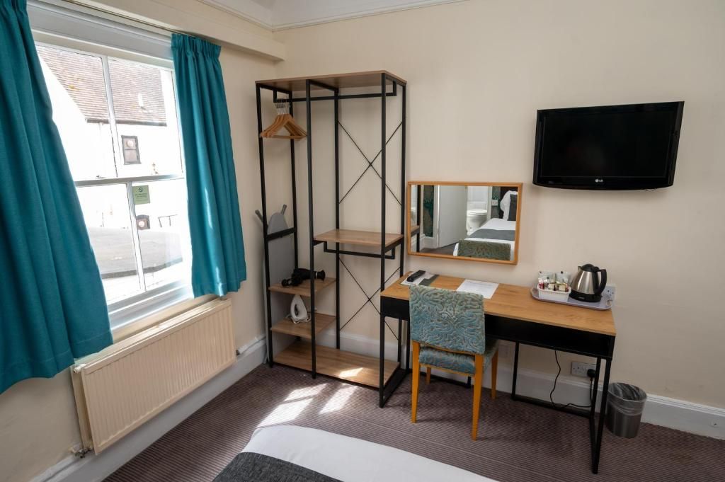A hotel room with a bed, a desk with a chair, a shelf unit, a television, and a window with curtains.