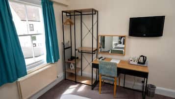 A hotel room with a bed, a desk with a chair, a shelf unit, a television, and a window with curtains.