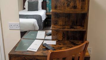 Un escritorio de madera con un espejo que refleja una cama y una ventana. Hay papeles, un control remoto y un menú sobre el escritorio.