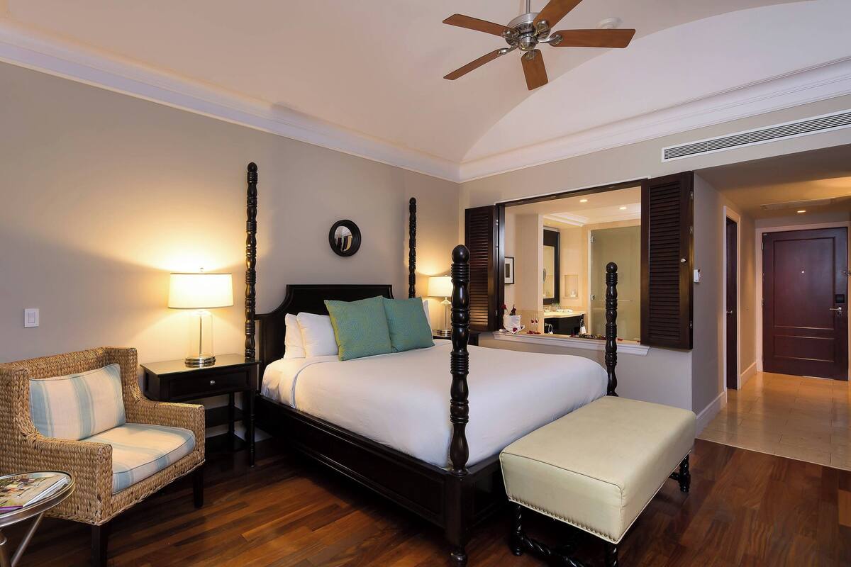 A four-poster bed with a wooden headboard, a bedside table with a lamp, a wicker armchair, and a bench.