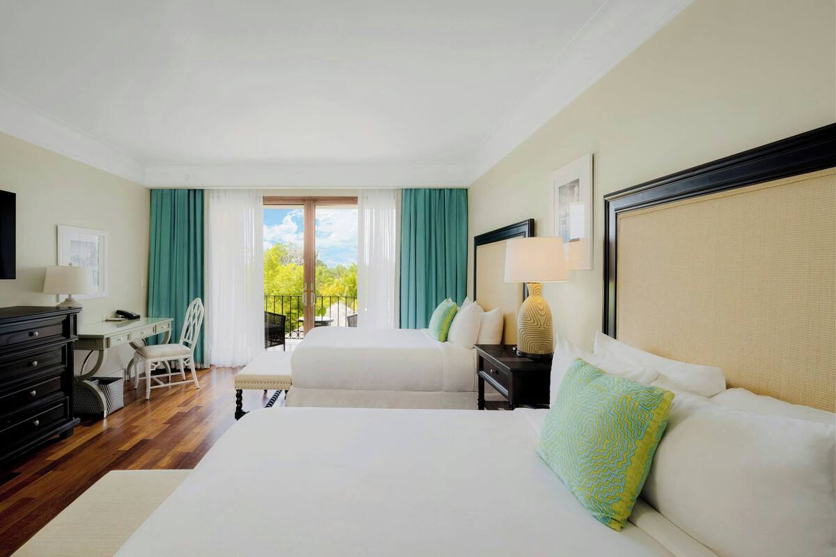A hotel room with two beds, a desk, a chair, and a view of the outdoors.