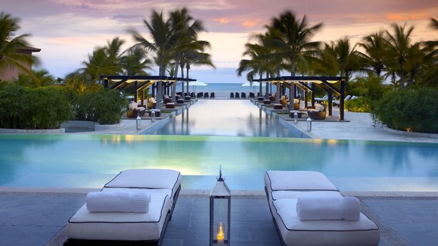 A poolside lounge area with two white cushioned chairs and a lantern, surrounded by palm trees and a view of the ocean at sunset. - The Buenaventura Golf & Beach Resort Panama, Autograph Collection (El Chiru)