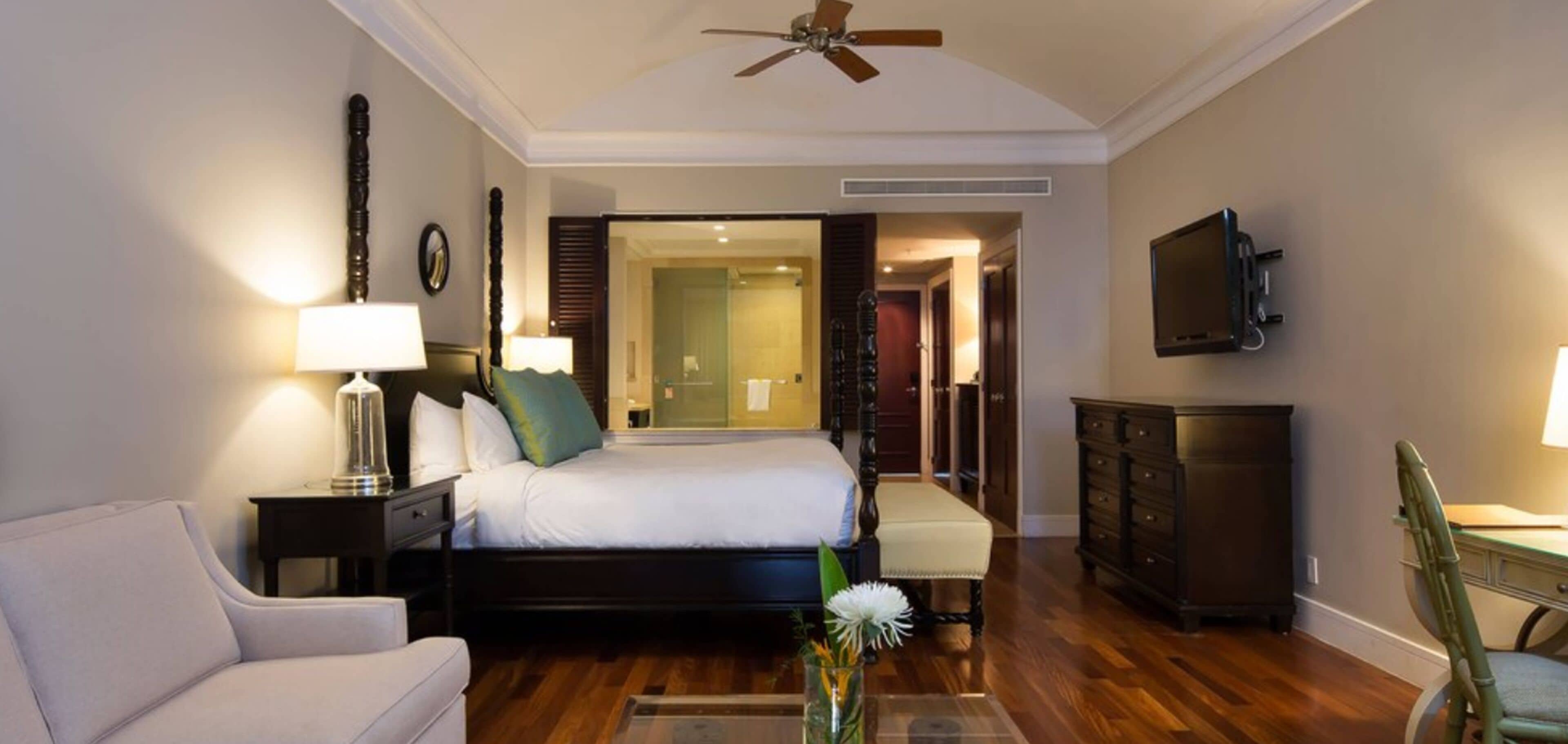 A hotel room with a bed, a desk, a chair, a TV, a ceiling fan, and a nightstand with a lamp.