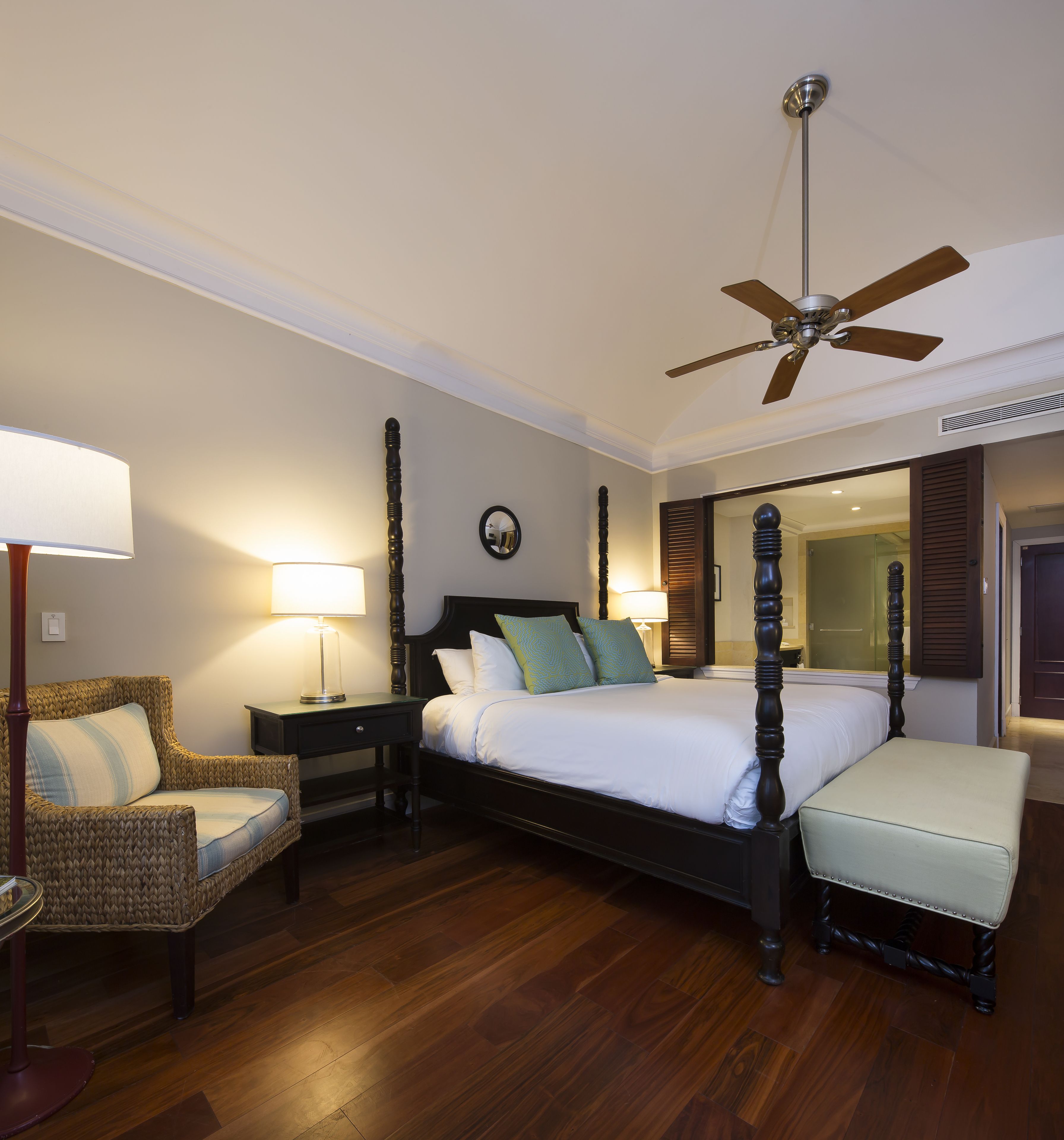 A hotel room with a four-poster bed, a chair, a bedside table, a ceiling fan, and a large mirror.