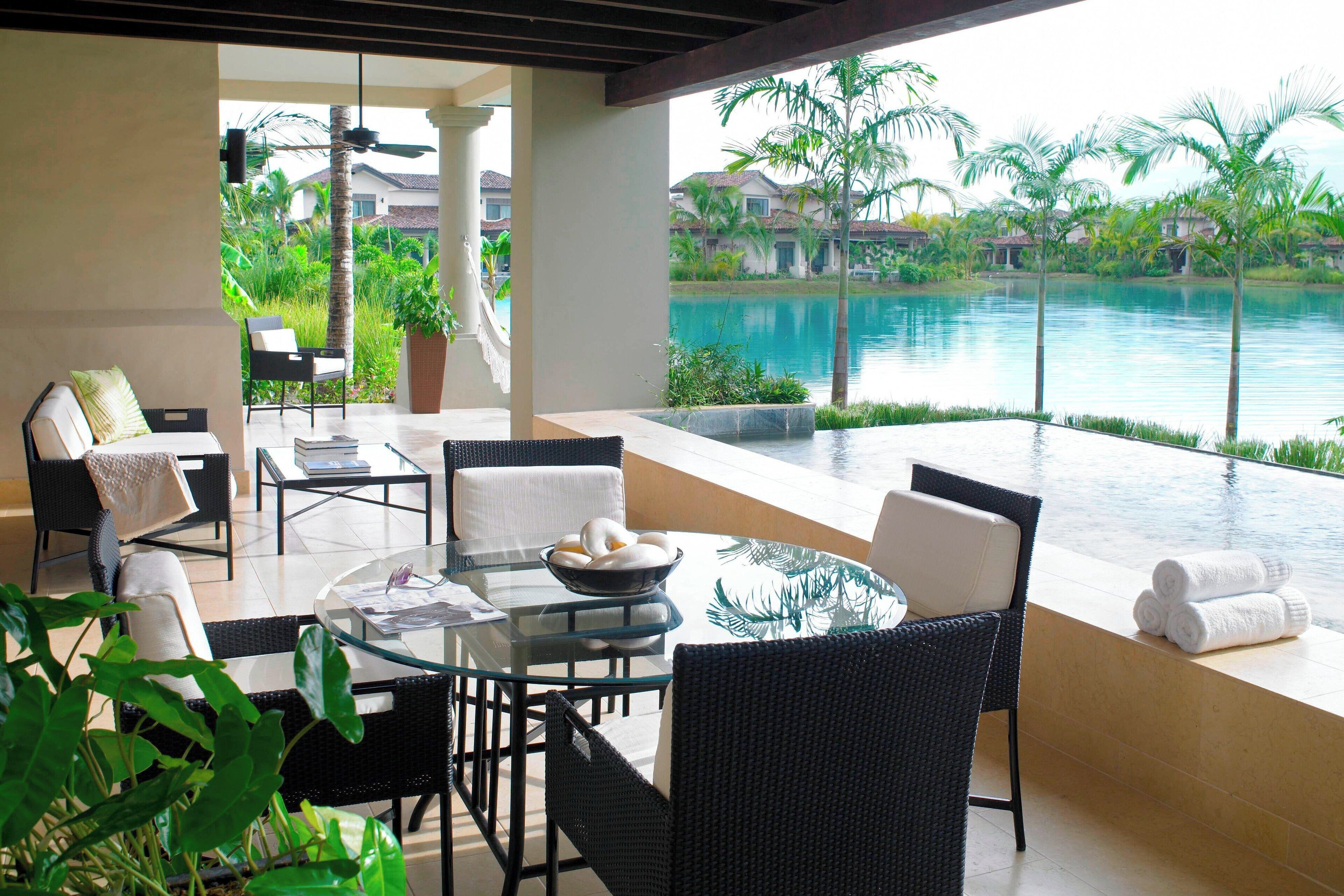 Un área de estar moderna al aire libre con mesa de vidrio y sillas de mimbre, con vistas a una piscina y abundante vegetación.