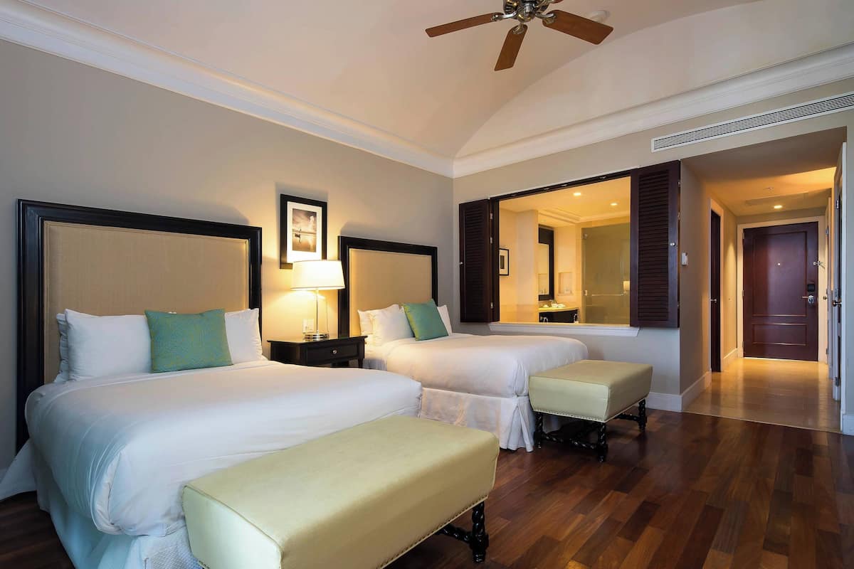 A hotel room with two beds, a bench, a ceiling fan, and a mirror.