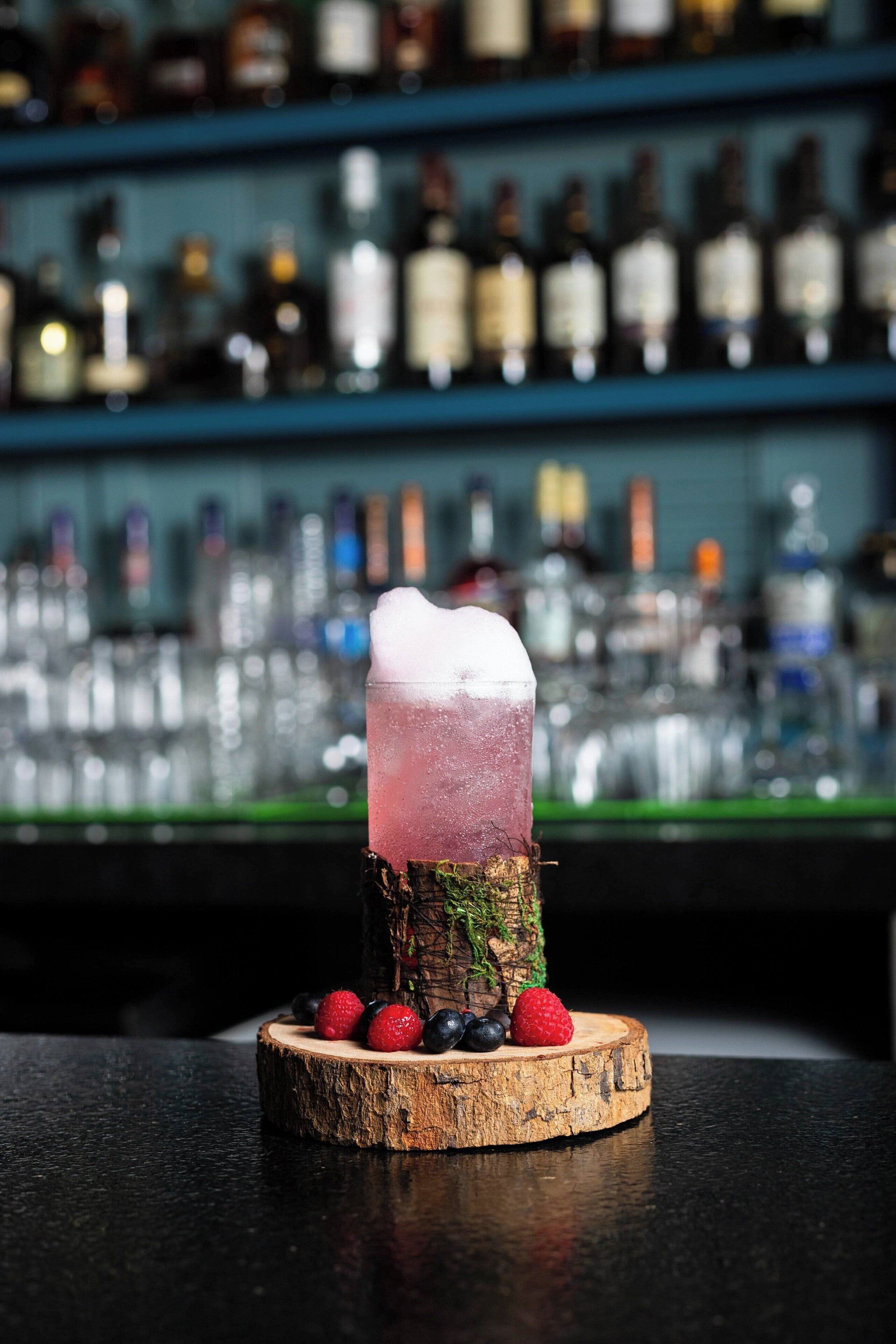 A cocktail on a wooden coaster with berries, set against a bar with shelves of bottles.