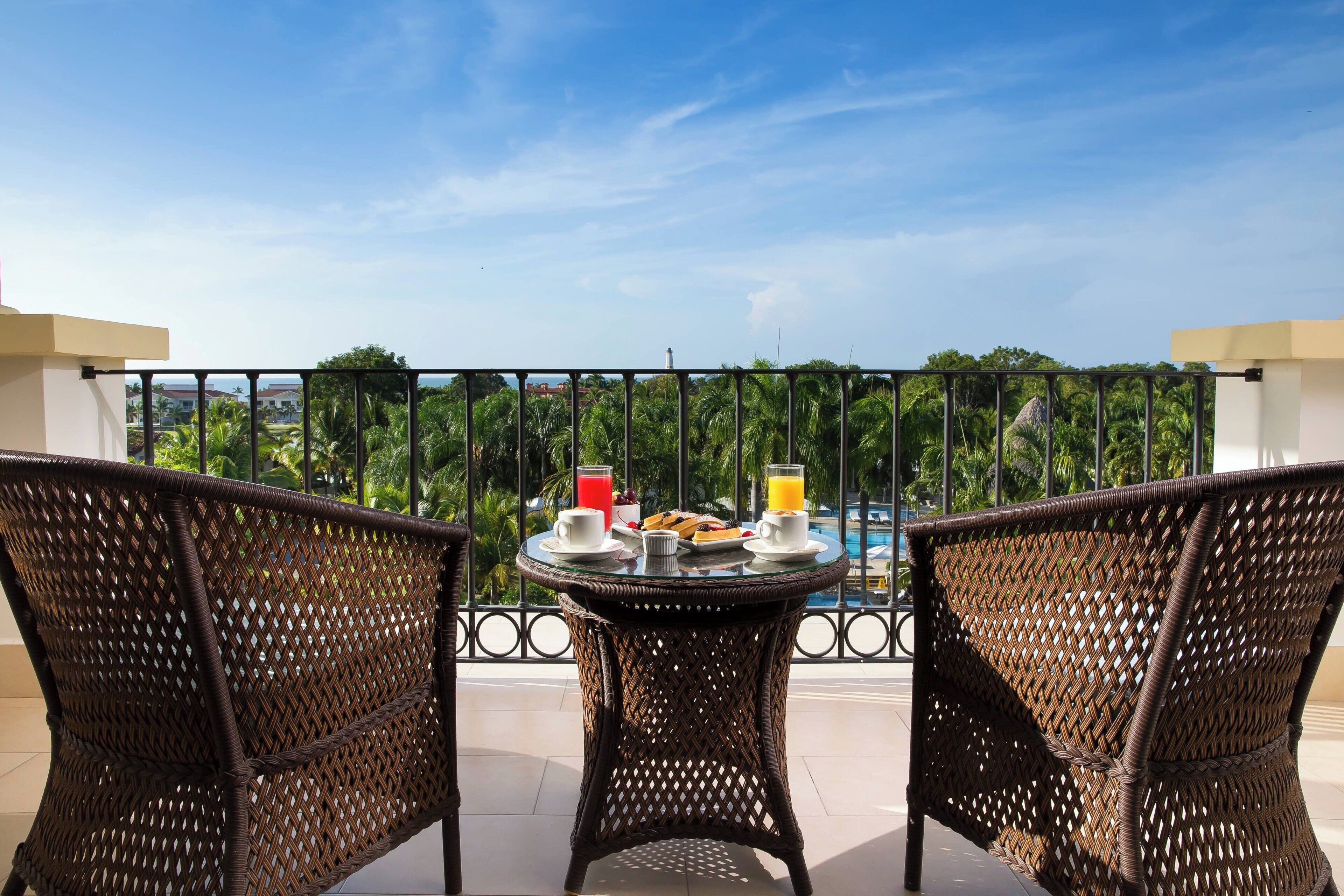 Un balcón con mobiliario de mimbre, una mesa con alimentos para el desayuno y vista a la piscina.