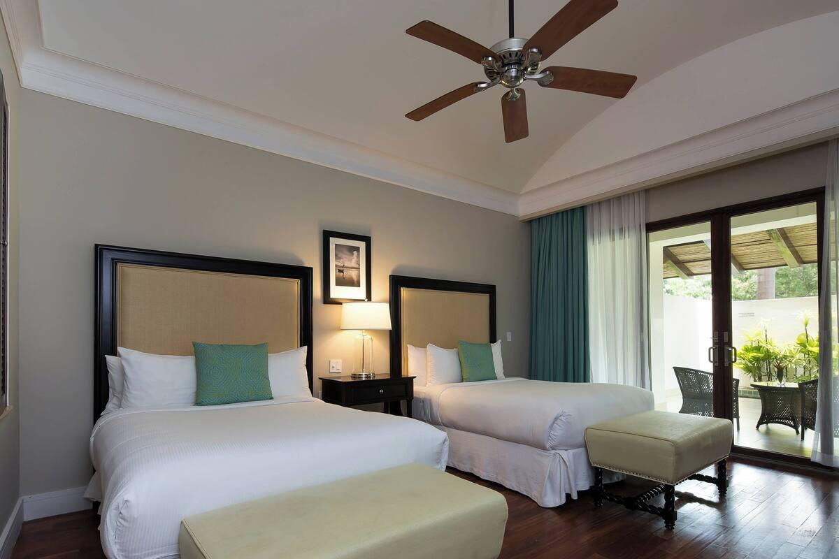 A hotel room with two beds, a ceiling fan, a window with curtains, and a view of the outdoors.