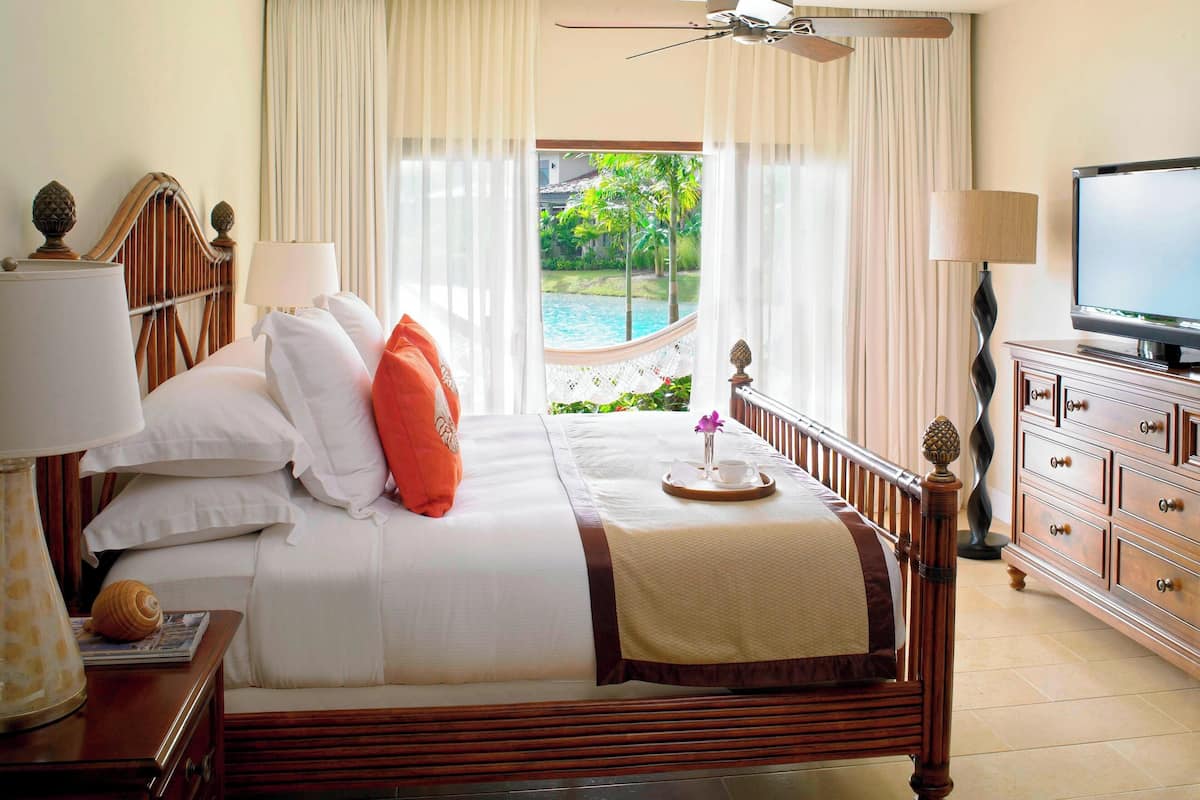 A hotel room with a bed, a television, a nightstand with a lamp, and a view of a swimming pool.