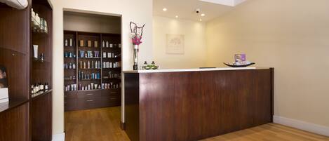 A reception area with a wooden reception desk, a wall-mounted shelf with toiletries, and a hallway leading to a door.