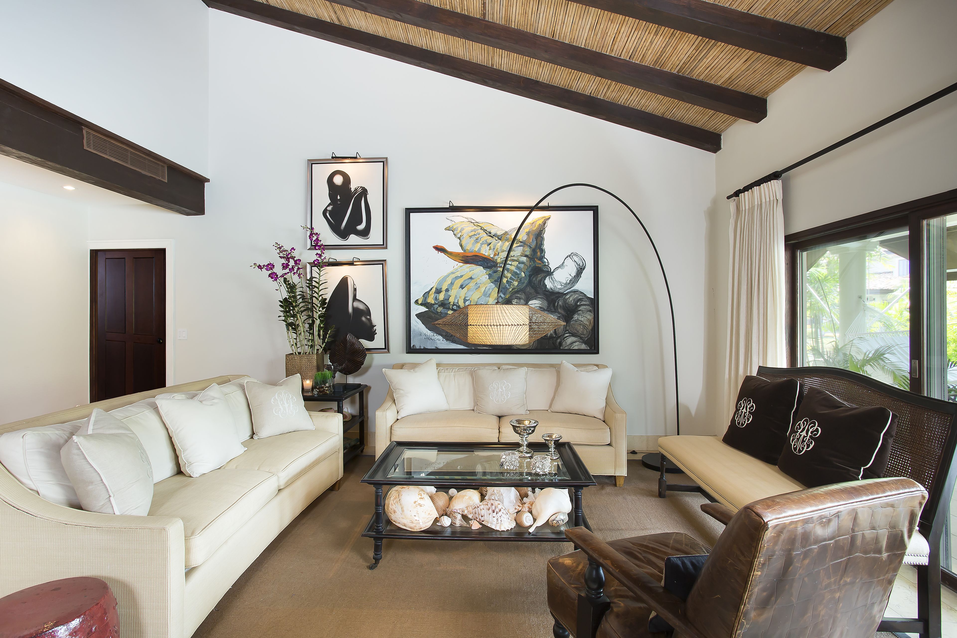 A living room with a beige sofa, a glass coffee table with decorative shells, a brown leather armchair, and a large framed artwork above the sofa.