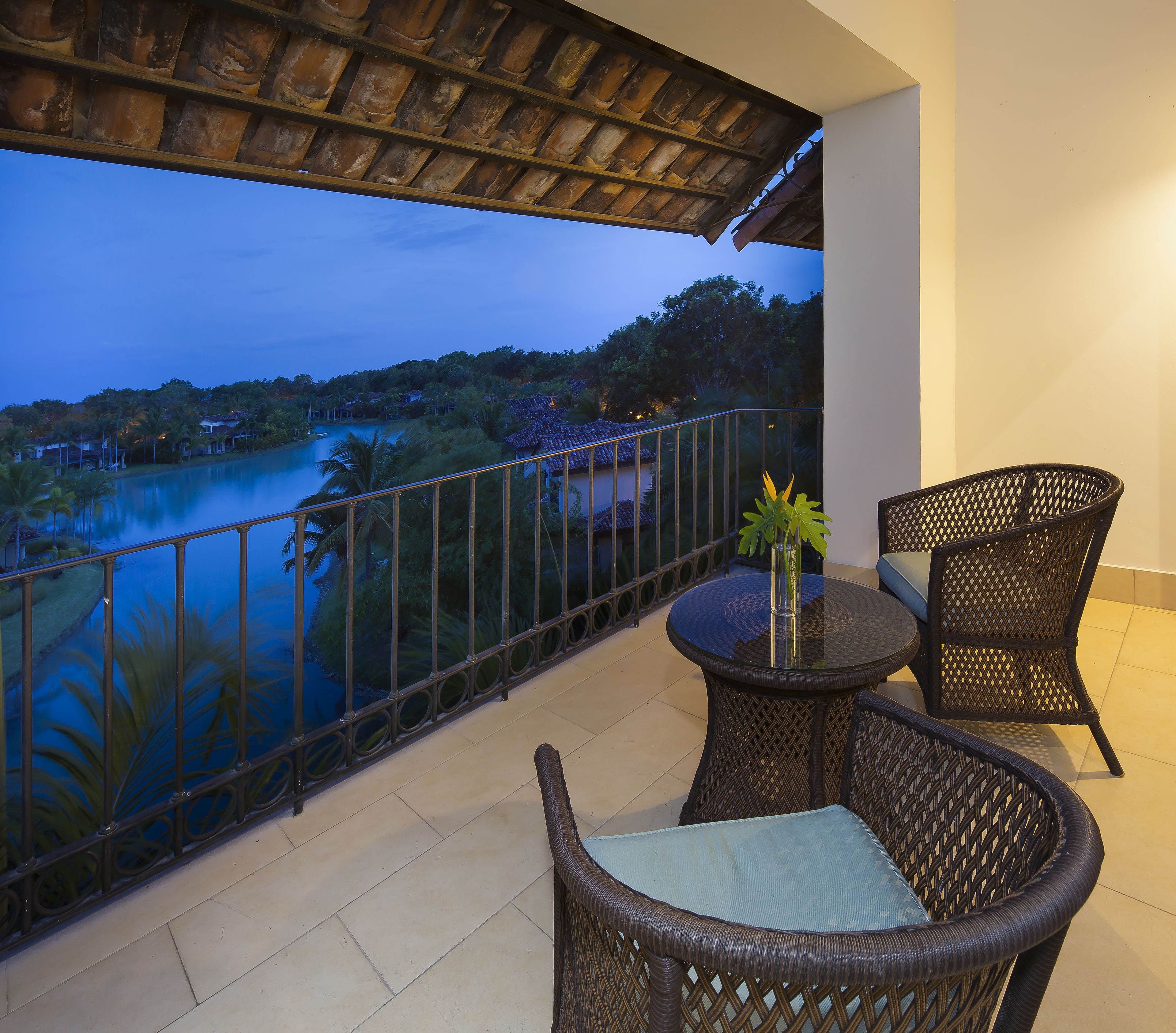 Un balcón con mobiliario de mimbre, una mesa de vidrio y vista al lago y árboles al anochecer.
