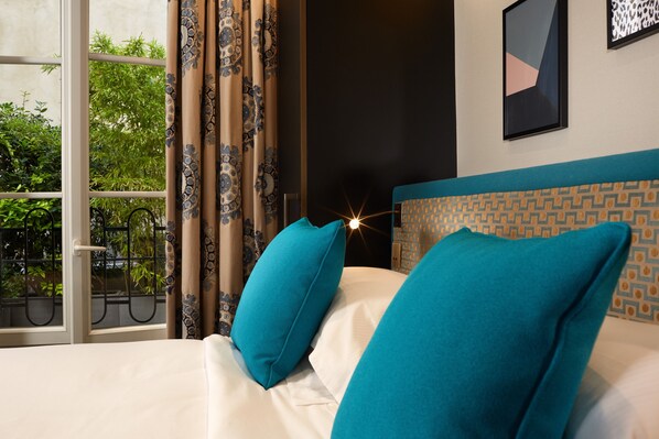 Una cama con almohadas color turquesa, un cabecero estampado y vistas al verdor a través de una ventana con cortinas decoradas.