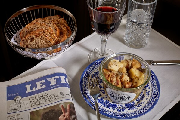 Une table avec un journal, un verre de vin rouge, un panier de pain et un bocal de nourriture.
