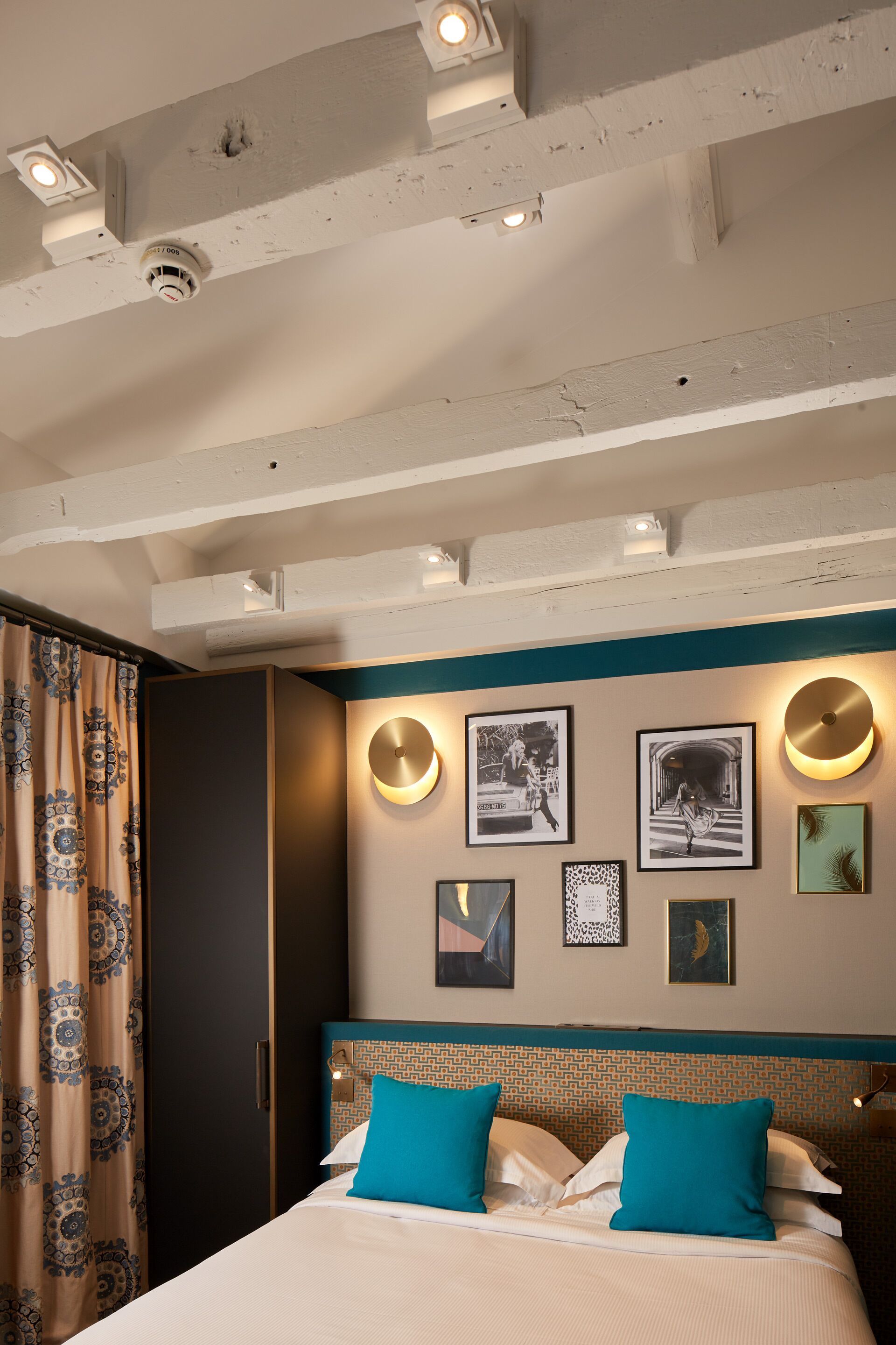 Habitación de hotel con cama, cabecera de diseño estampado, pared con cuadros enmarcados e iluminación empotrada en el techo.