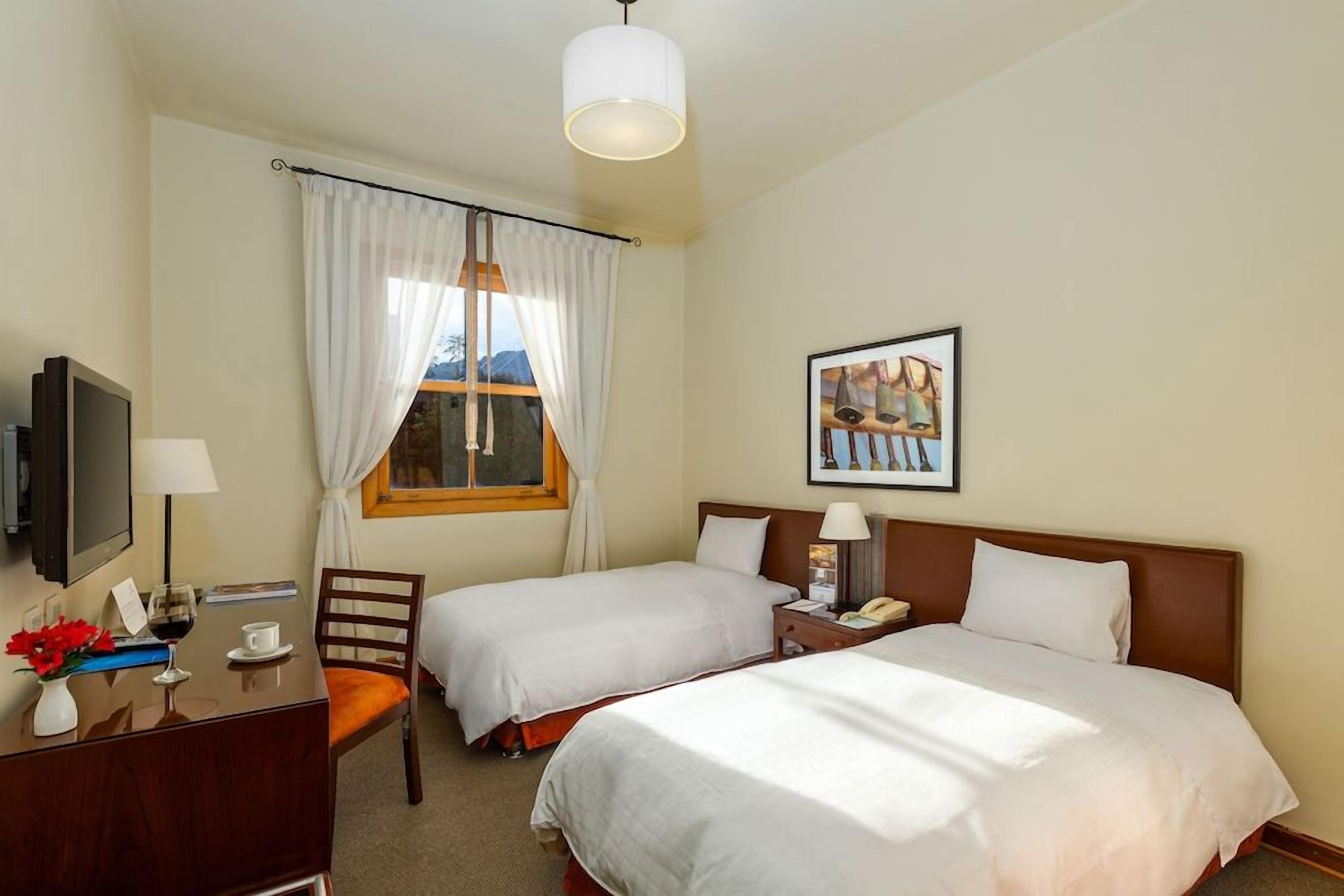 A hotel room with two beds, a desk with a chair, a TV, and a window with curtains.