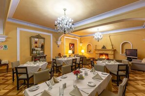 A dining area with multiple tables set for guests, chandeliers, a fireplace, and a television.