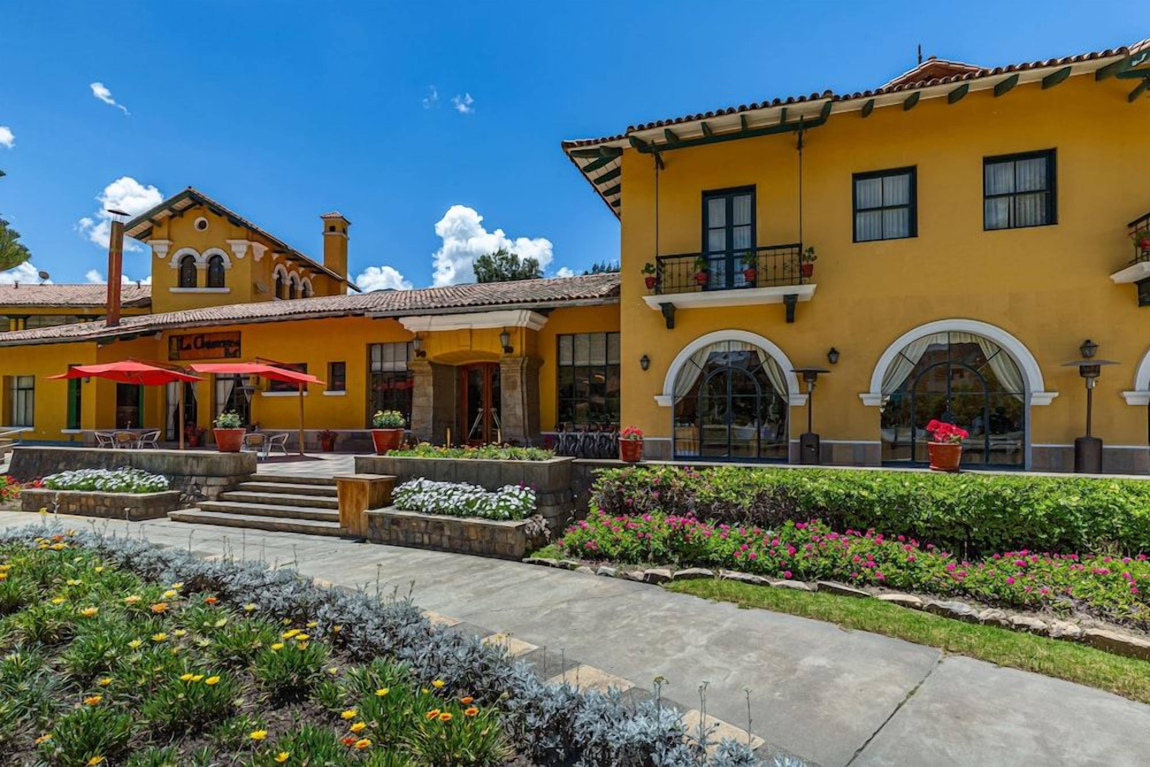 Un edificio amarillo con un patio, mesas al aire libre y macizos de flores.