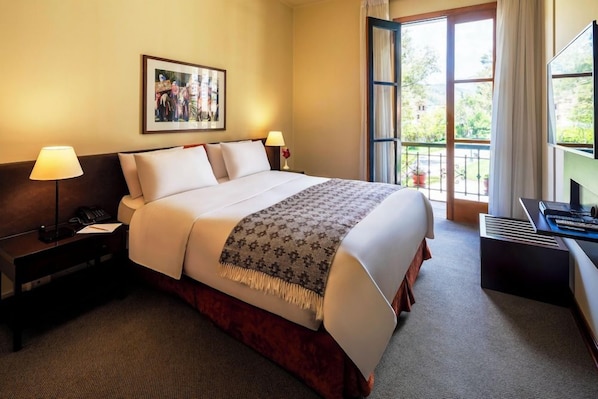 A hotel room with a bed, bedside lamps, a desk with a computer, and a window with a view of greenery.