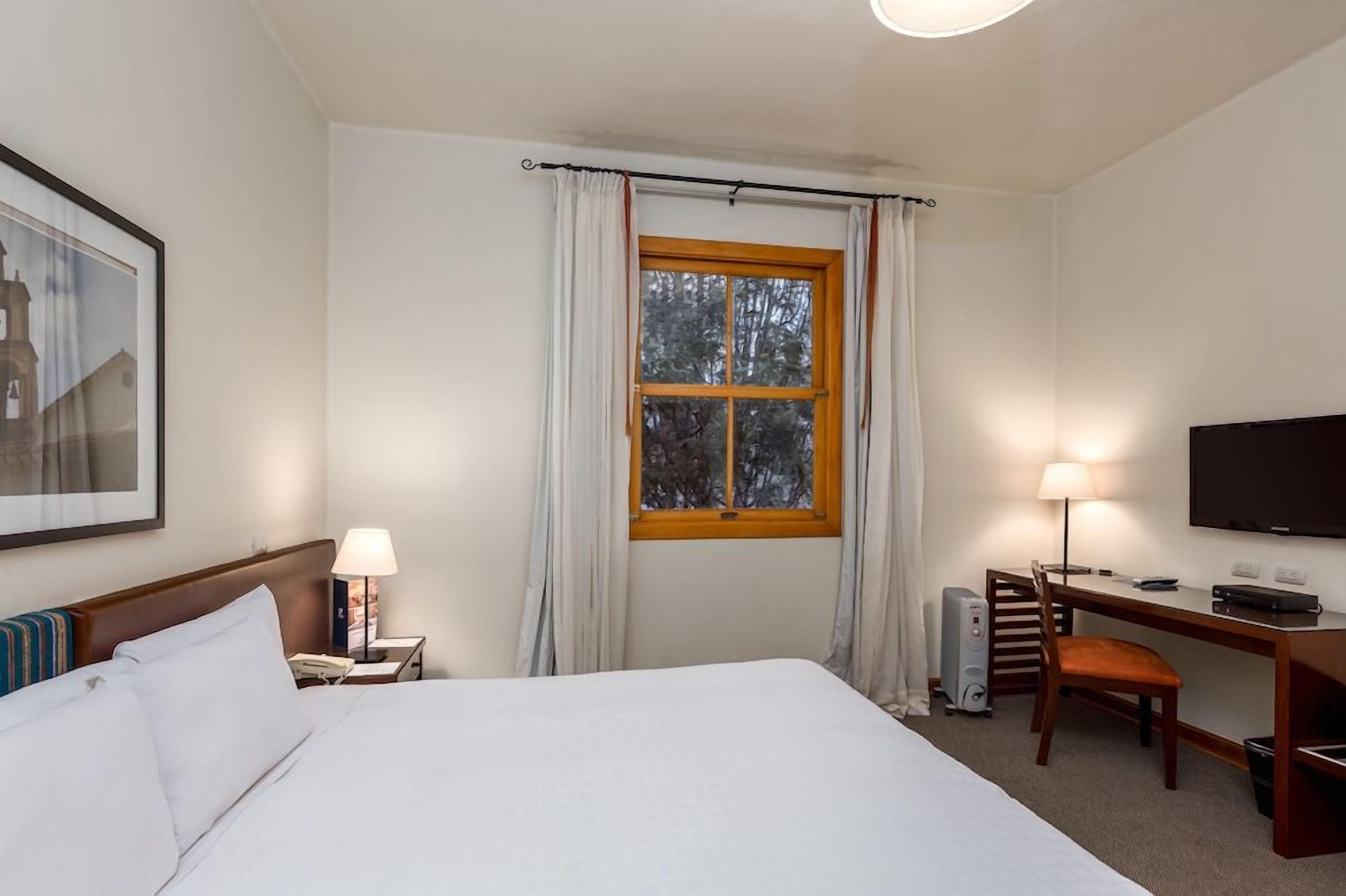 Habitación de hotel con cama, escritorio, silla, televisor y ventana con cortinas.