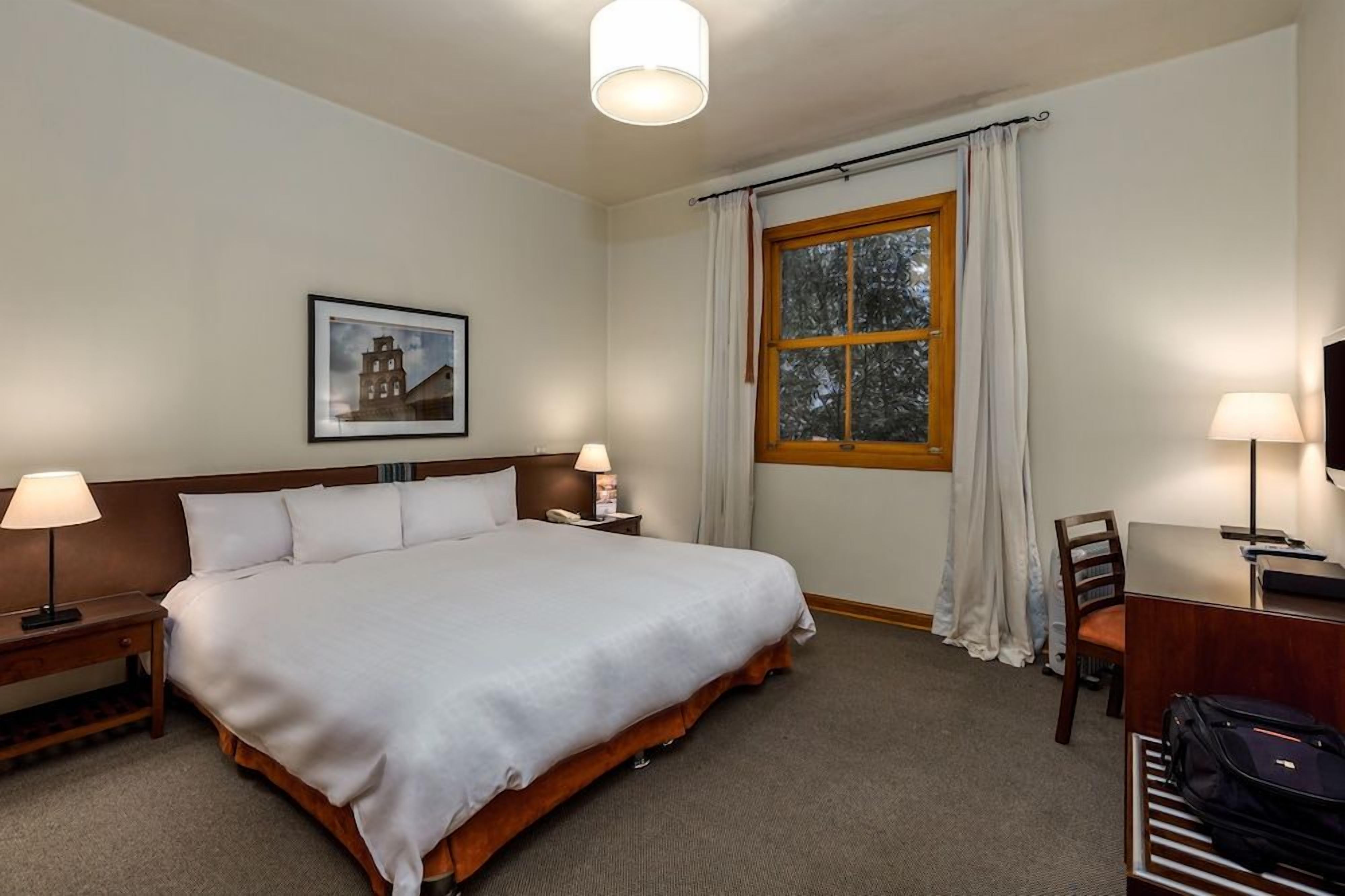 Habitación de hotel con cama, lámparas de noche, escritorio con silla, ventana con cortinas e imagen enmarcada en la pared.