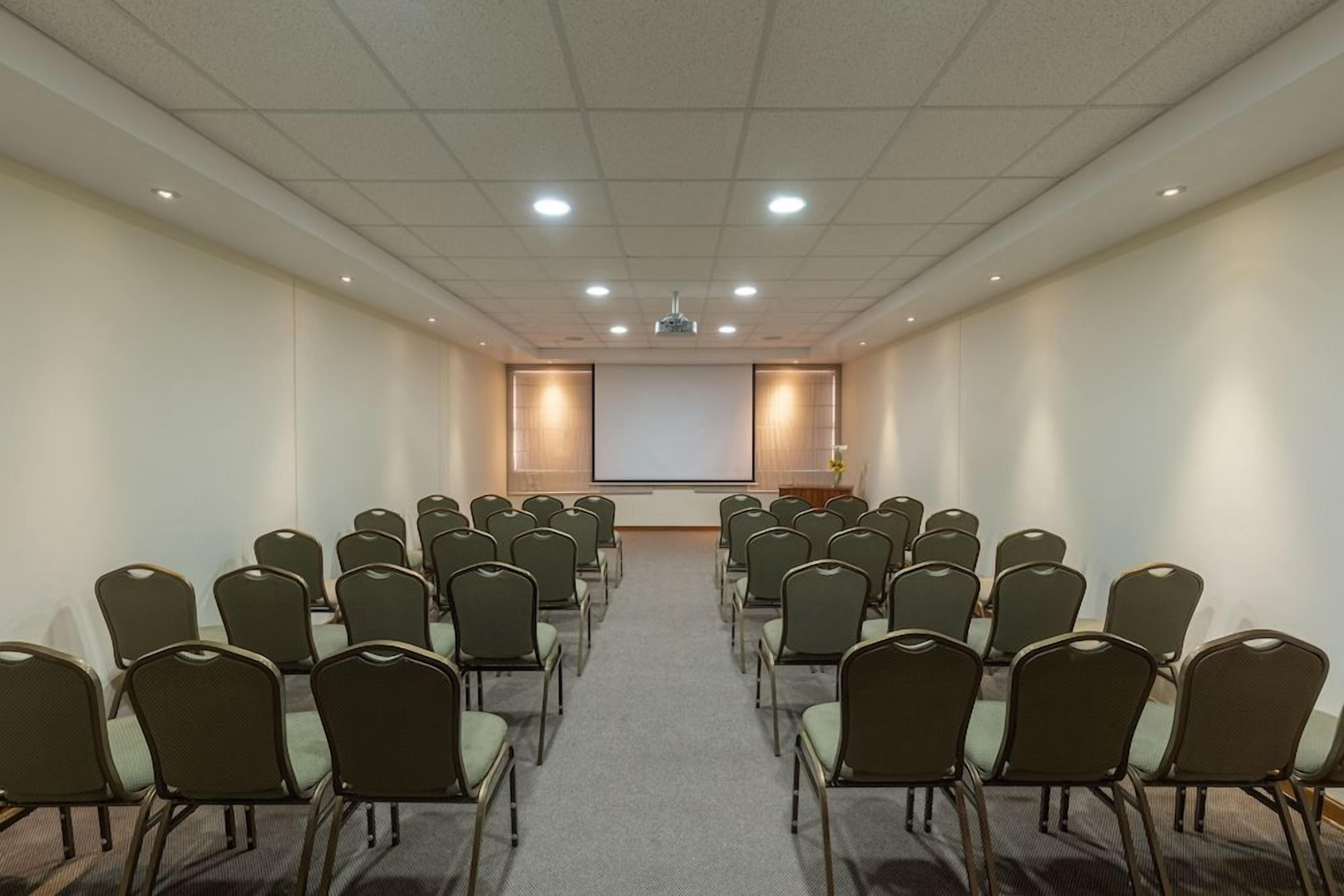 A conference room with rows of chairs facing a projection screen, a podium, and a carpeted floor.
