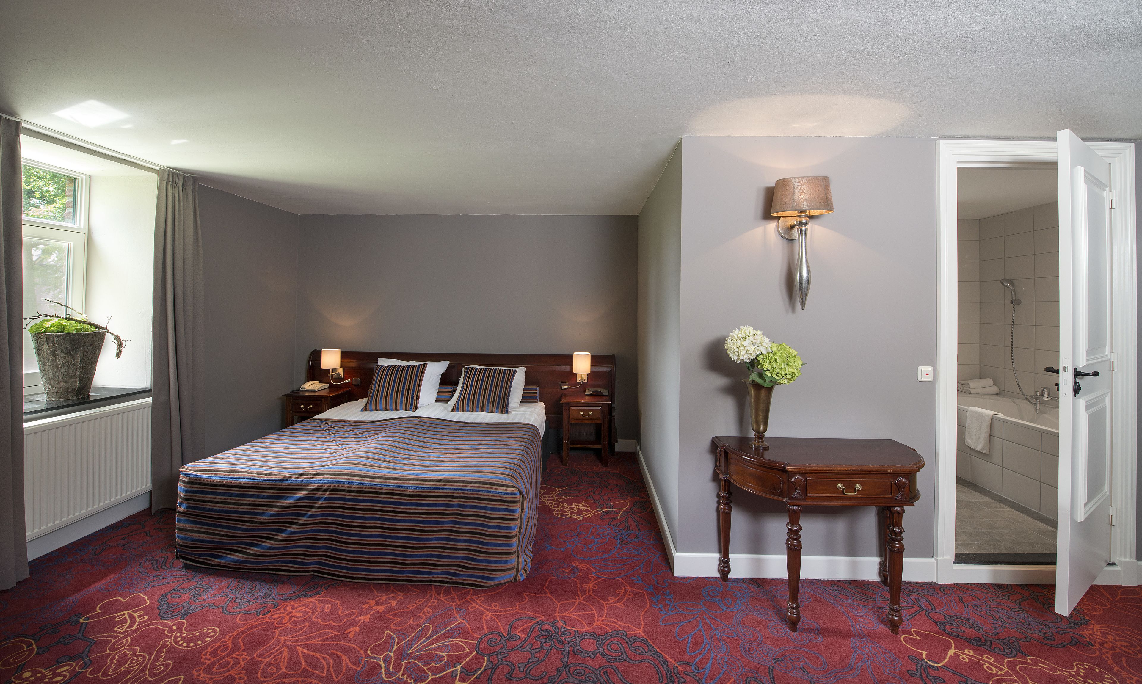 Habitación de hotel con una cama grande, mesitas de noche con lámparas, una planta en maceta, un jarrón con flores y un baño visible a través de una puerta abierta.
