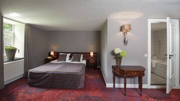 Habitación de hotel con una cama grande, mesitas de noche con lámparas, una planta en maceta, un jarrón con flores y un baño visible a través de una puerta abierta.