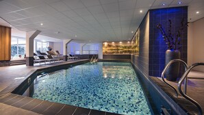 Una piscina con piso de baldosas, sillas de descanso y una pared decorativa con un paisaje natural.